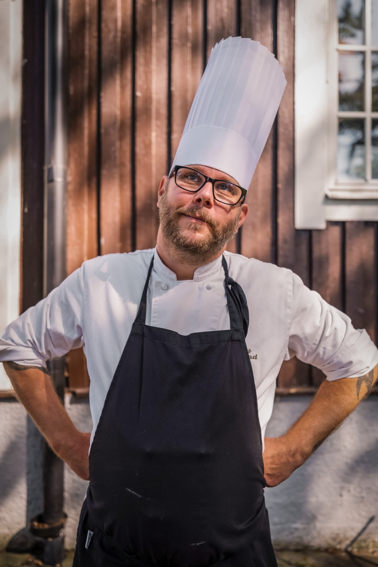Barkland utomhus i vit kockjacka och hög kockmössa framför brunt trähus